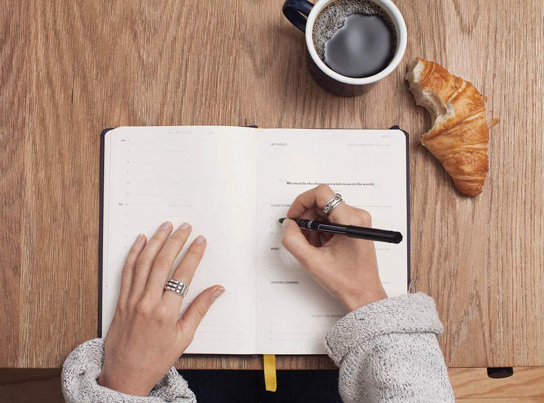 overhead view of hands writing in notebook, coffee and croissant on table